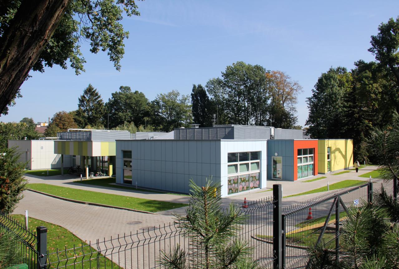 Nowoczesny, parterowy budynek o modułowej konstrukcji z płaskim dachem i kolorowymi elementami fasady, otoczony zielenią i ogrodzeniem.