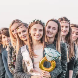 Grupa kobiet w wiankach na głowach, ustawiona w rzędzie, z przodu panna młoda z szarfą i balonem w kształcie pierścionka zaręczynowego.