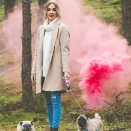 Kobieta w płaszczu spaceruje z dwoma mopsami w lesie z różową smugą dymu w tle.