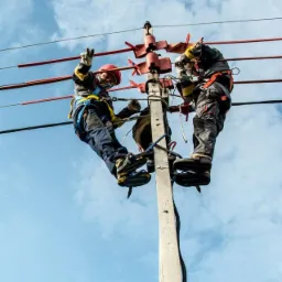 Dwóch elektryków w kaskach i uprzężach pracuje na słupie energetycznym, jeden z nich pokazuje znak zwycięstwa na tle błękitnego nieba.