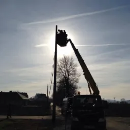 Pracownicy na podnośniku koszowym przy słupie energetycznym na tle zachodzącego słońca, widoczny samochód serwisowy i linia energetyczna.