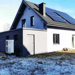 Nowoczesny dom jednorodzinny z panelami słonecznymi na dachu i zamontowaną pompą ciepła Panasonic na zewnątrz budynku, otoczony trawnikiem z resztkami śniegu.