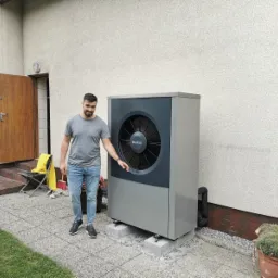 Mężczyzna prezentuje zamontowaną pompę ciepła Buderus na betonowych podstawach przy ścianie budynku z fakturą baranka, widoczne elementy instalacji oraz otwarte drzwi wejściowe.