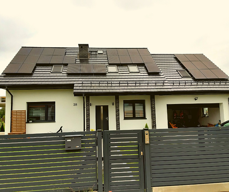 Dom jednorodzinny z czarną dachówką i panelami słonecznymi na dachu, widoczne okna dachowe i numer domu 38, ogrodzenie panelowe w kolorze grafitowym.