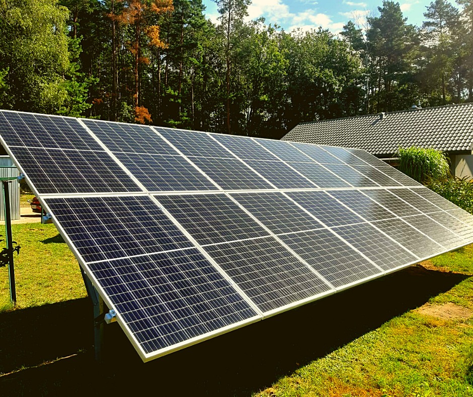 Instalacja paneli słonecznych w Knurowie, widok na moduły fotowoltaiczne na tle zielonej trawy, drzew i budynku z ciemną dachówką.