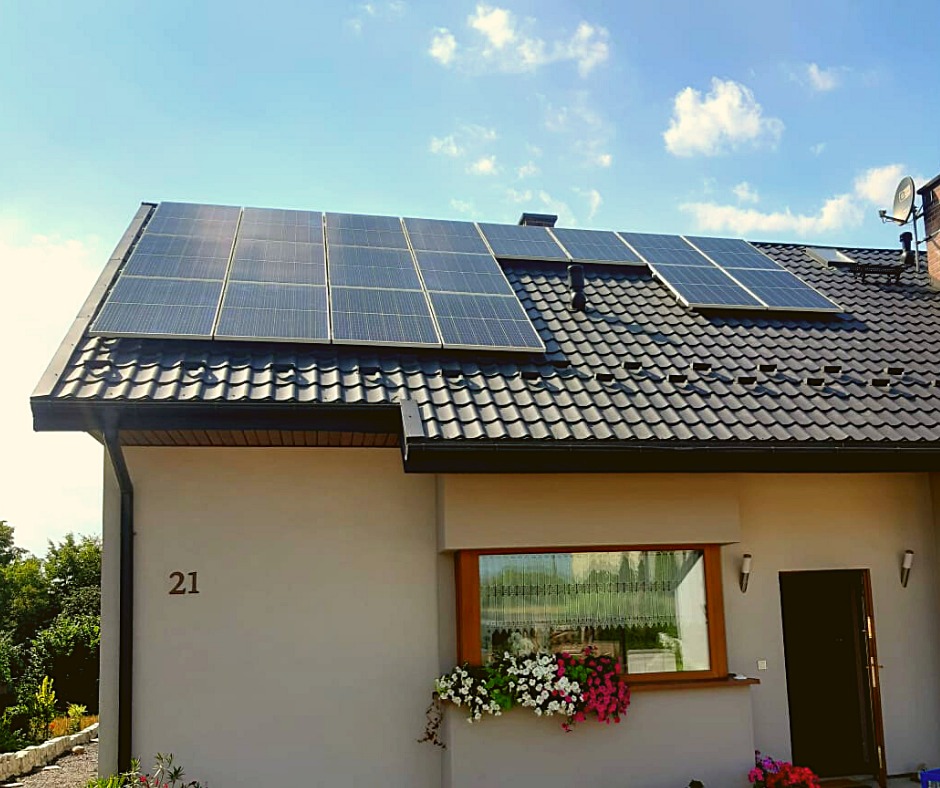 Panele słoneczne na dachu domu jednorodzinnego z ciemną dachówką, widok na elewację z numerem 21 i oknem ozdobionym kwiatami.