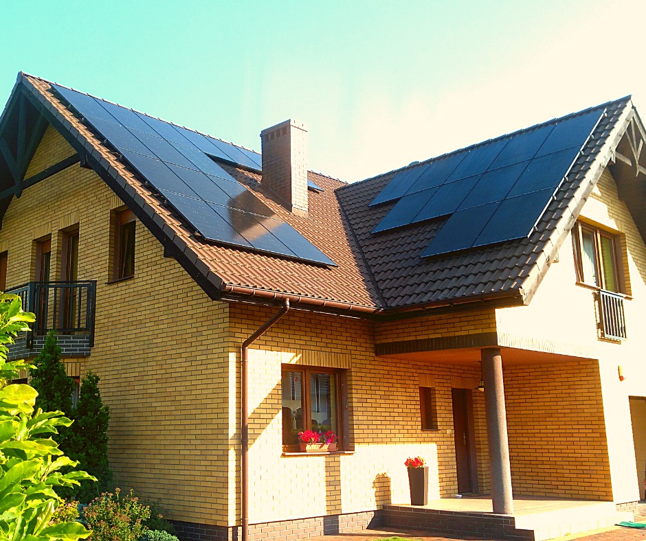 Dom jednorodzinny z żółtej cegły z brązowym dachem i panelami fotowoltaicznymi na połaciach dachu, widok z poziomu ogrodu z zielenią.