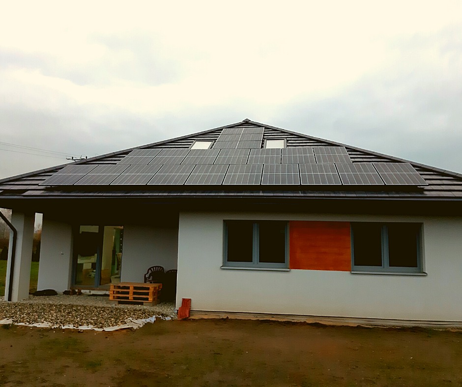 Dom jednorodzinny z panelami słonecznymi na dachu, widoczne okno dachowe i tymczasowe zabezpieczenie okna na elewacji bocznej, teren wokół domu w trakcie zagospodarowania.