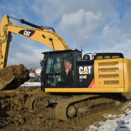 Żółta koparka CAT 324E nabiera ziemię z wykopu, widoczny operator w kabinie, w tle budynki i fragmenty ośnieżonego terenu.