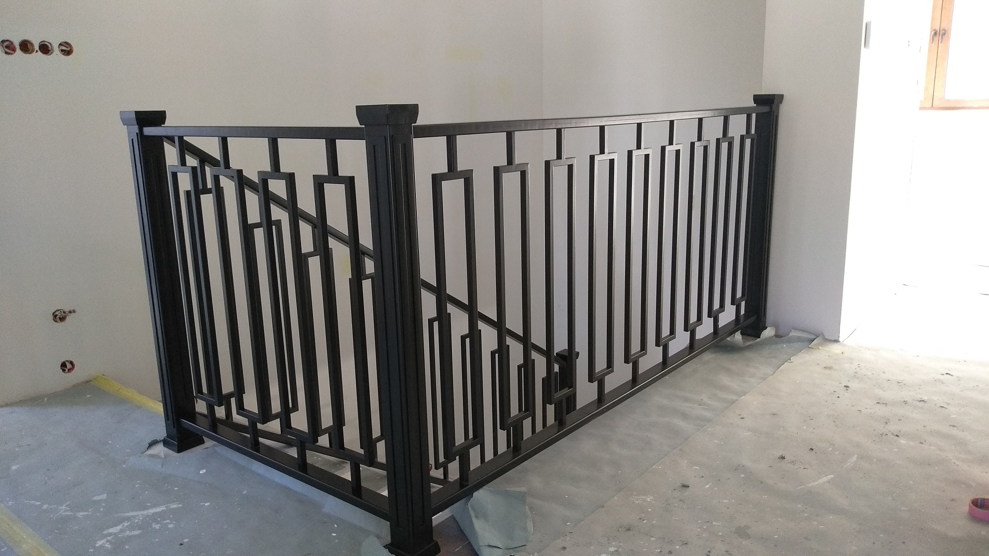 Czarna, metalowa balustrada schodowa o geometrycznym wzorze w surowym wnętrzu, widoczne przygotowania do wykończenia ścian i podłogi.