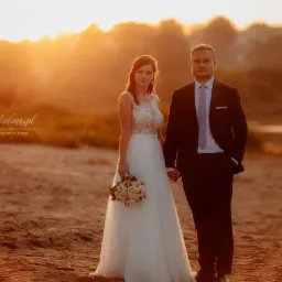 Para młoda trzymająca się za ręce na plaży o zachodzie słońca, panna młoda w białej sukni z bukietem, pan młody w czarnym garniturze, ciepłe światło zachodzącego słońca.