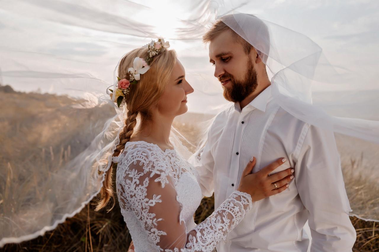 Para młoda w plenerze: panna młoda w koronkowej sukni i wianku z kwiatów spogląda na pana młodego w białej koszuli, oboje owiani delikatnym woalem na tle łąki w ciepłym świetle.