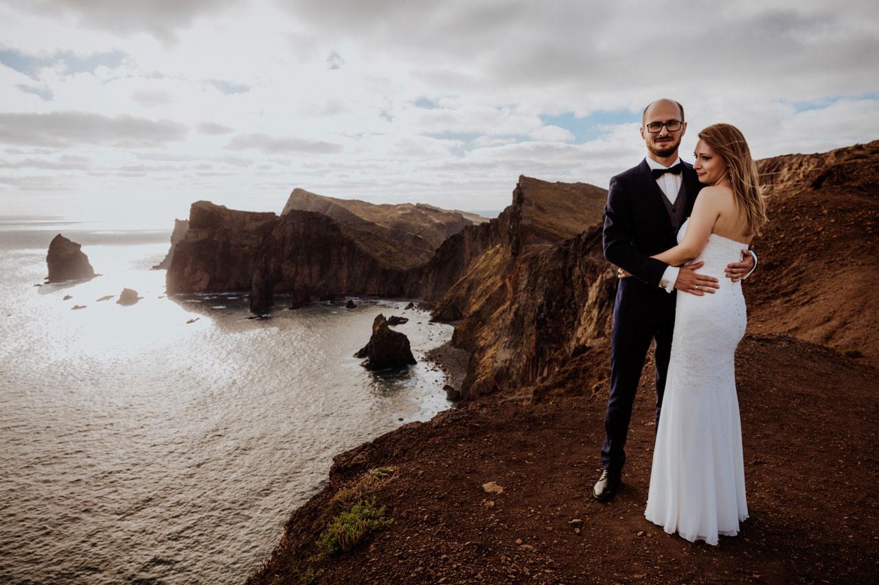 Para młoda w objęciach na szczycie klifu z widokiem na ocean i skaliste wybrzeże w tle, on w granatowym garniturze z muszką, ona w białej sukni ślubnej bez ramiączek.