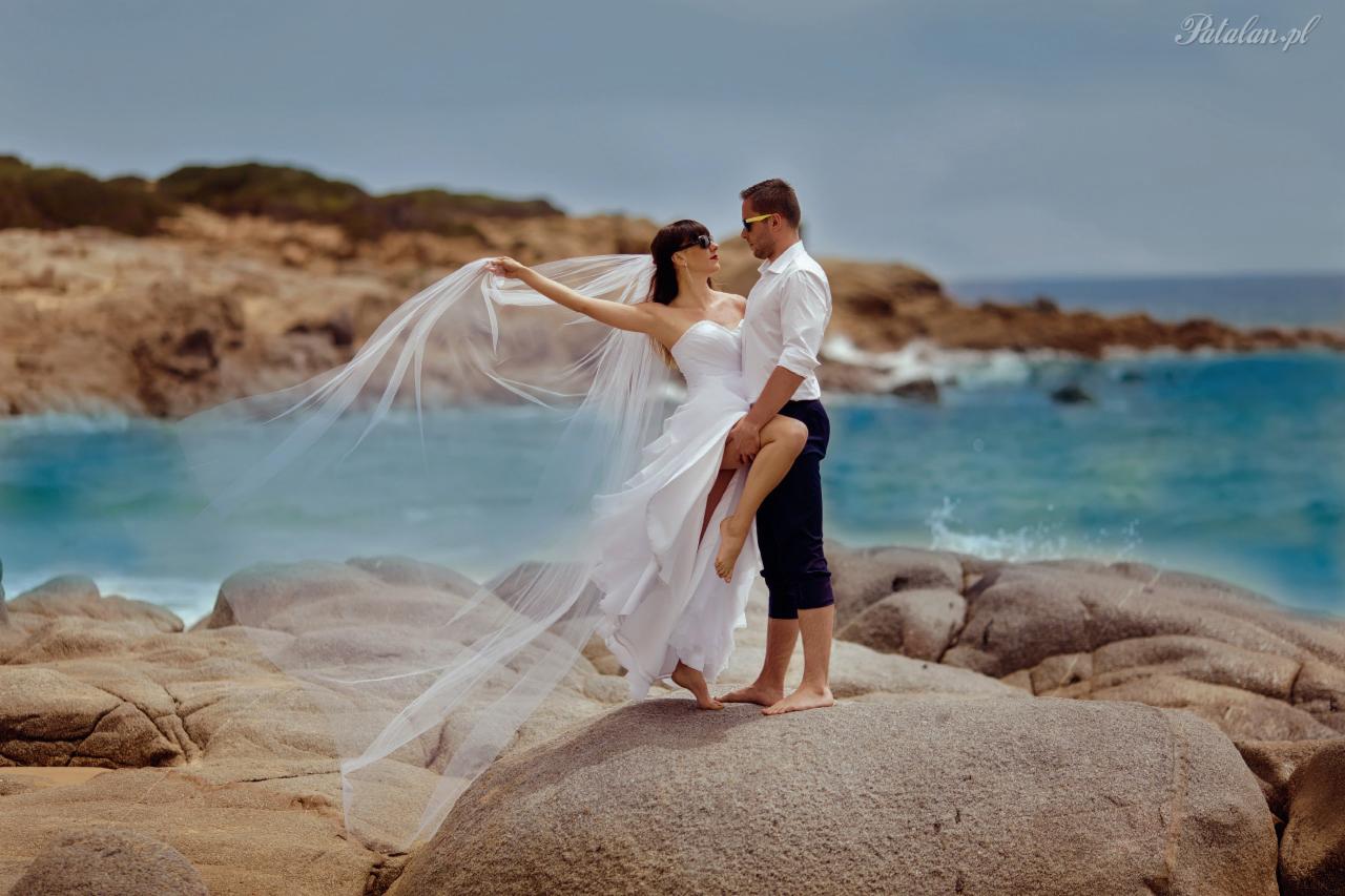 Para młoda w okularach przeciwsłonecznych pozuje na skalistej plaży; panna młoda w zwiewnej sukni i welonie, podtrzymywana przez pana młodego, z morzem i brzegiem w tle.