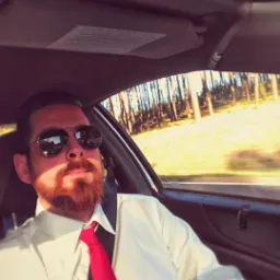 Mężczyzna z brodą i okularami przeciwsłonecznymi w samochodzie, ubrany w białą koszulę i czerwony krawat, zapinający pas bezpieczeństwa podczas podróży.