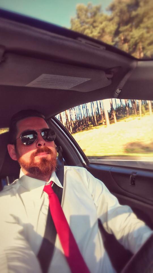 Mężczyzna z brodą i okularami przeciwsłonecznymi w samochodzie, ubrany w białą koszulę i czerwony krawat, zapinający pas bezpieczeństwa podczas podróży.