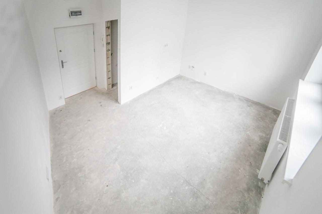 Surowe wnętrze w trakcie remontu, widoczne białe, świeżo pomalowane ściany, betonowa posadzka, grzejnik i drzwi wejściowe, z widocznymi śladami prac wykończeniowych.