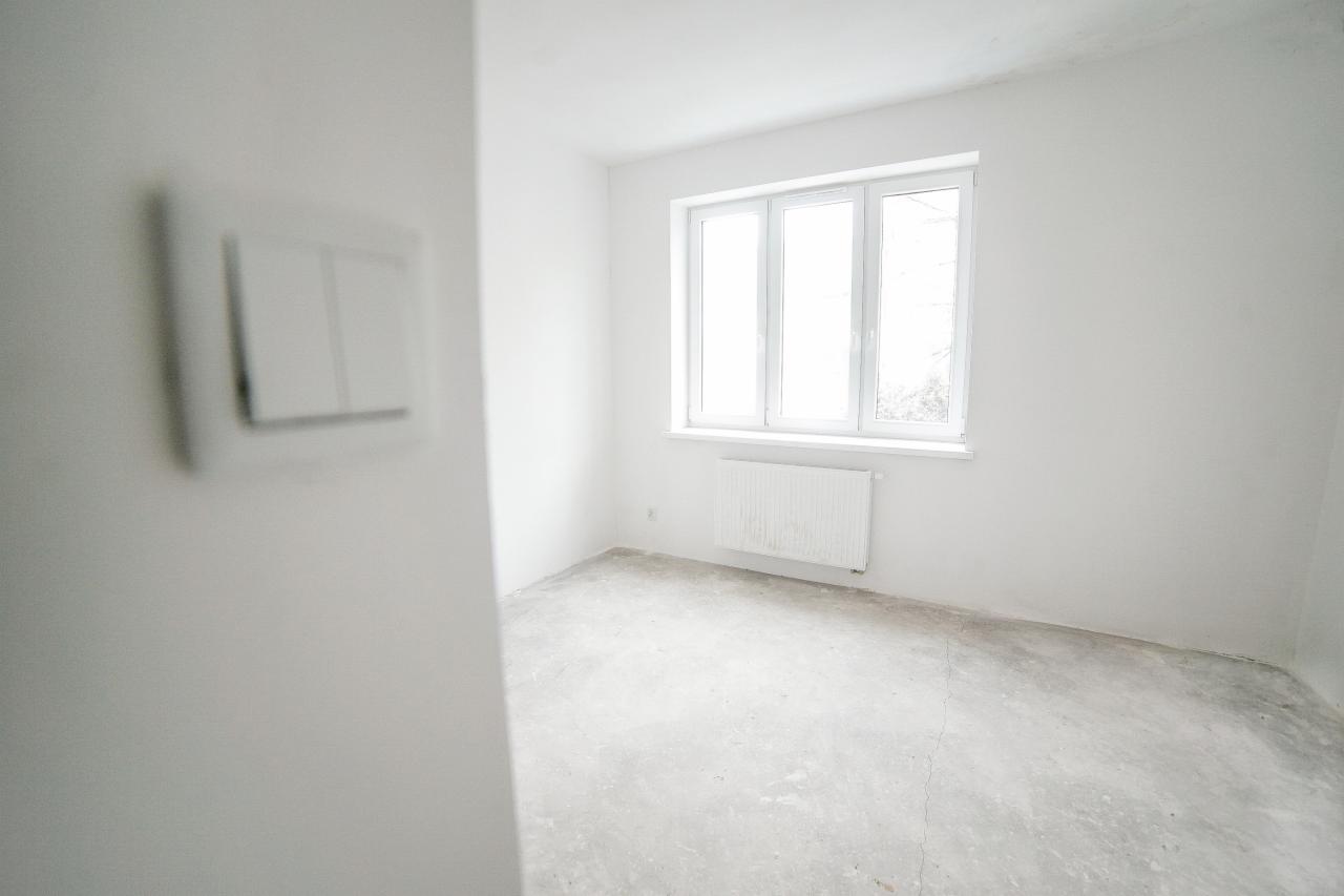 Pusty, biały pokój z oknem, grzejnikiem i surowym betonowym podłożem, widoczne przełączniki światła na ścianie.