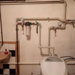 Instalacja wodna z filtrem, manometrami i bojlerem w pomieszczeniu z umywalką i płytkami w szachownicę, widoczne rury miedziane i plastikowe.