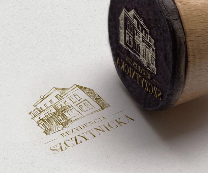 Złoty stempel z odciśniętym szkicem budynku i napisem 'Rezydencja Szczytnicka' na białym papierze, obok widoczny drewniany uchwyt stempla.