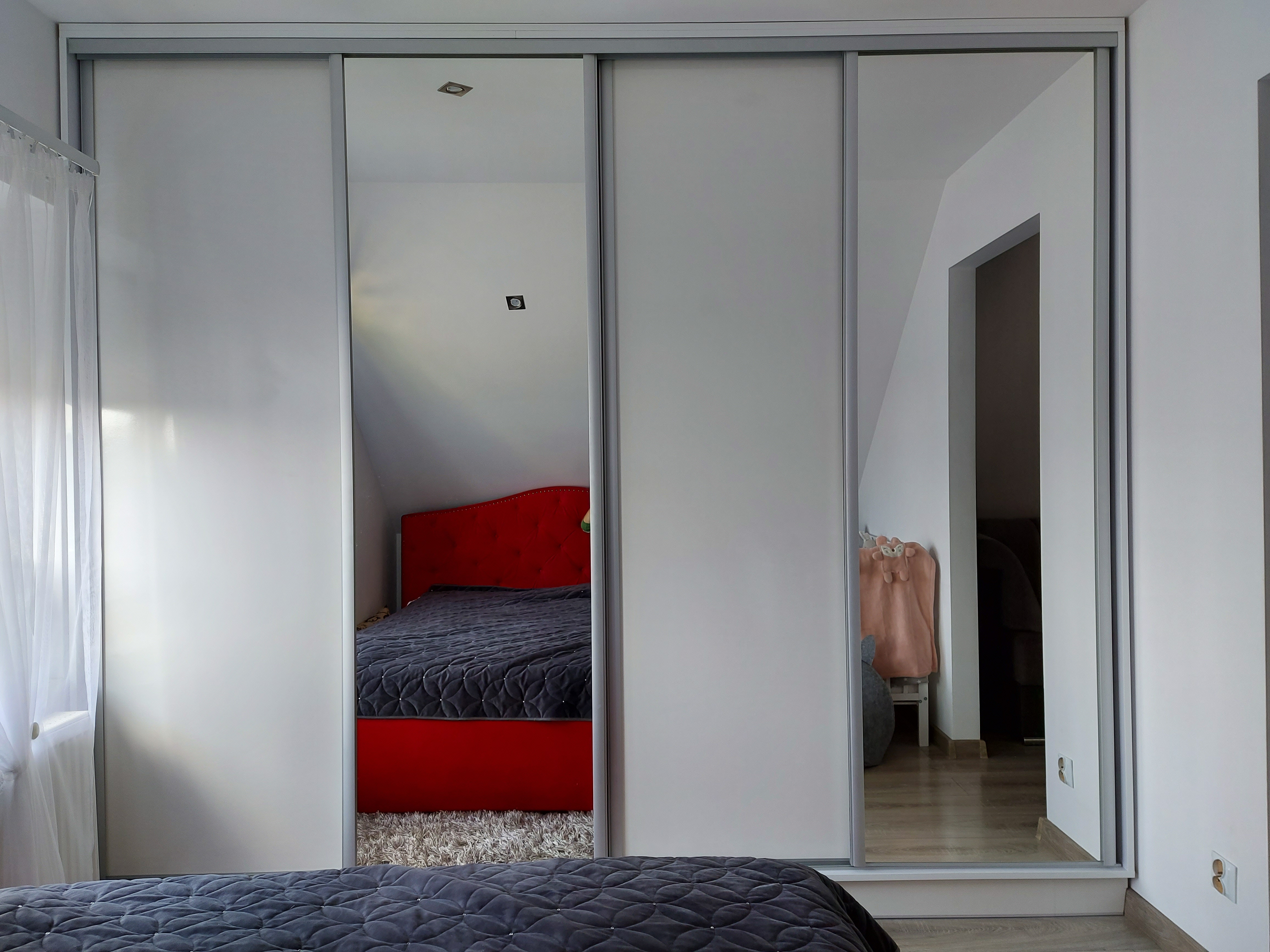 Biała szafa przesuwna z lustrami w sypialni na poddaszu, odbijająca czerwone wezgłowie łóżka i pikowaną narzutę.