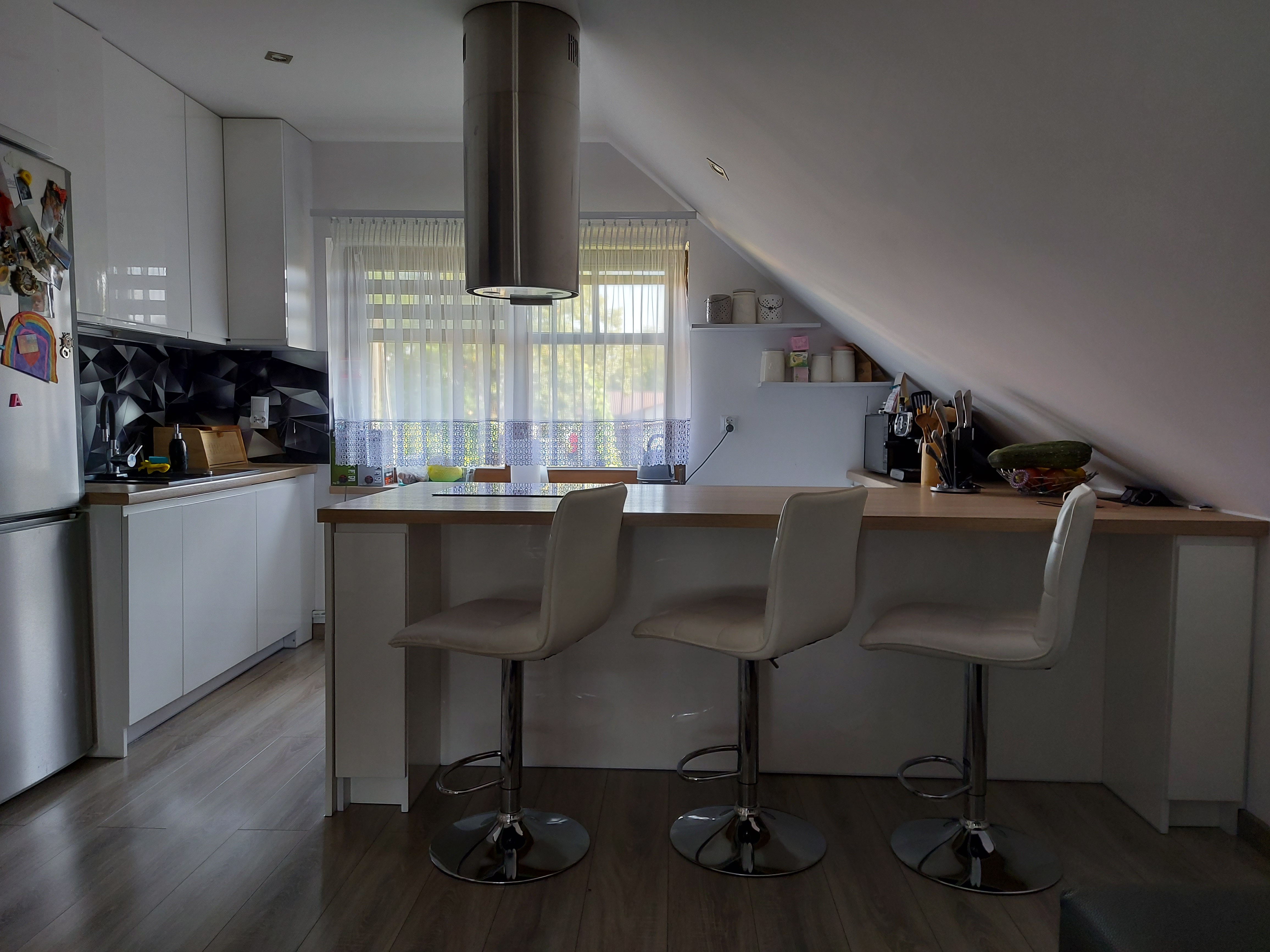 Kuchnia w skosie na poddaszu z białymi meblami, wyspą z trzema krzesłami barowymi i srebrnym okrągłym okapem.