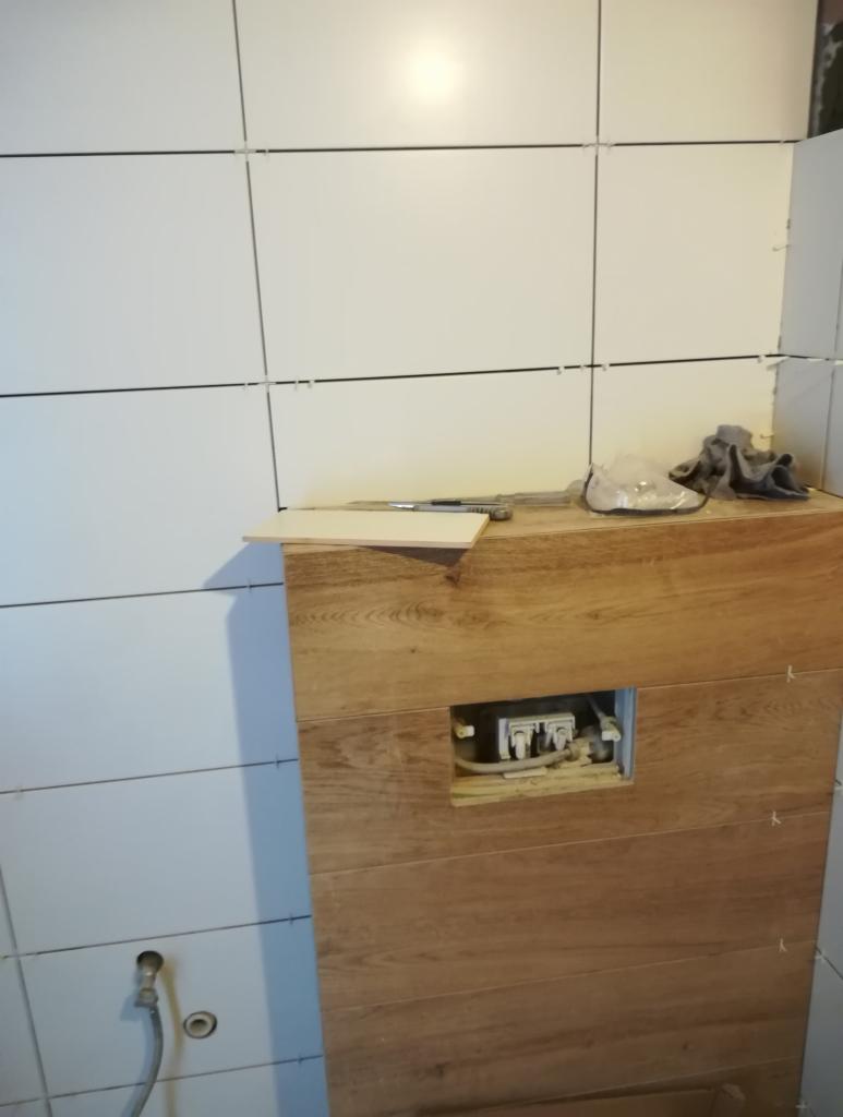 Montaż stelaża podtynkowego WC z drewnopodobną obudową, widoczne białe płytki na ścianie z krzyżykami dystansowymi i narzędzia na blacie