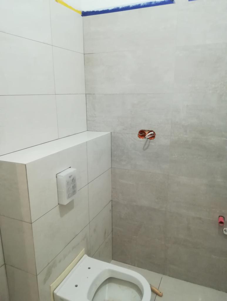 Niedokończona łazienka z szarymi płytkami, widoczny stelaż podtynkowy WC, niebieska taśma malarska przy suficie i wystające kable elektryczne.