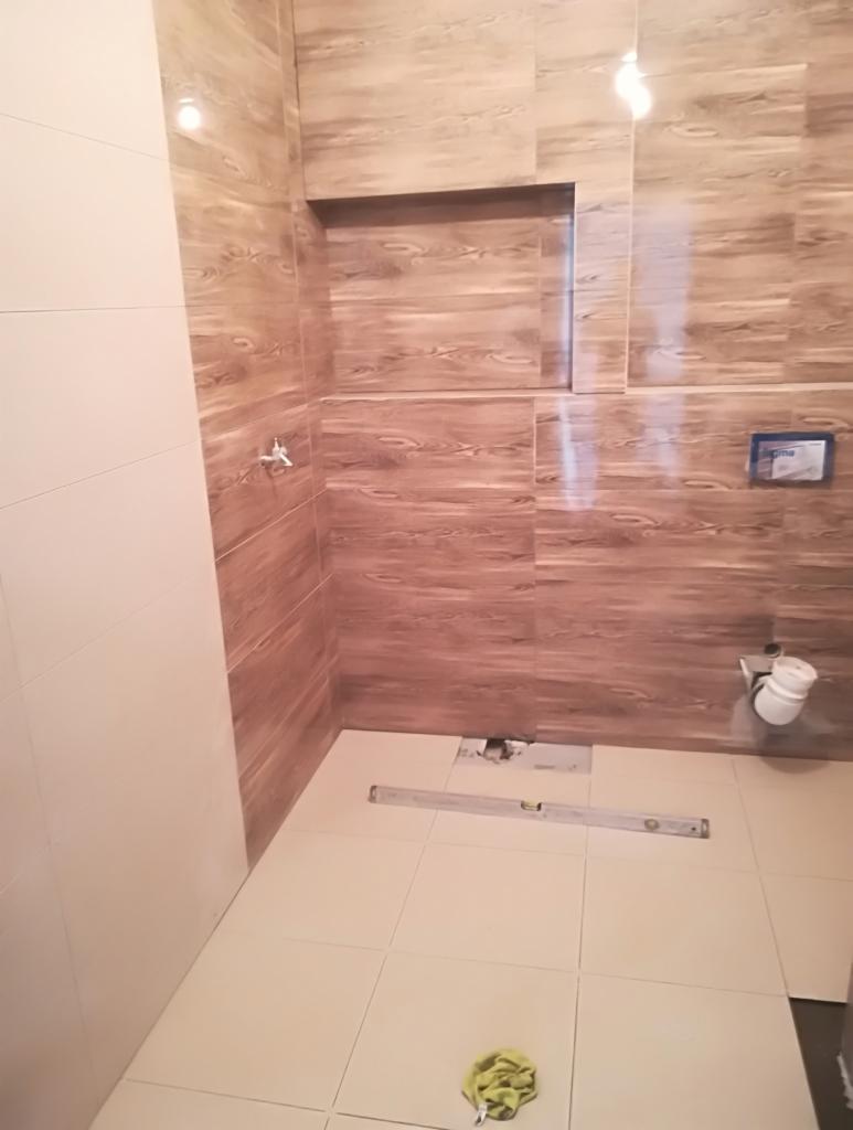 Łazienka w trakcie wykańczania: ściany wyłożone płytkami imitującymi drewno, wnęka w ścianie, częściowo położone płytki podłogowe, poziomica na podłodze, widoczny fragment instalacji toaletowej.