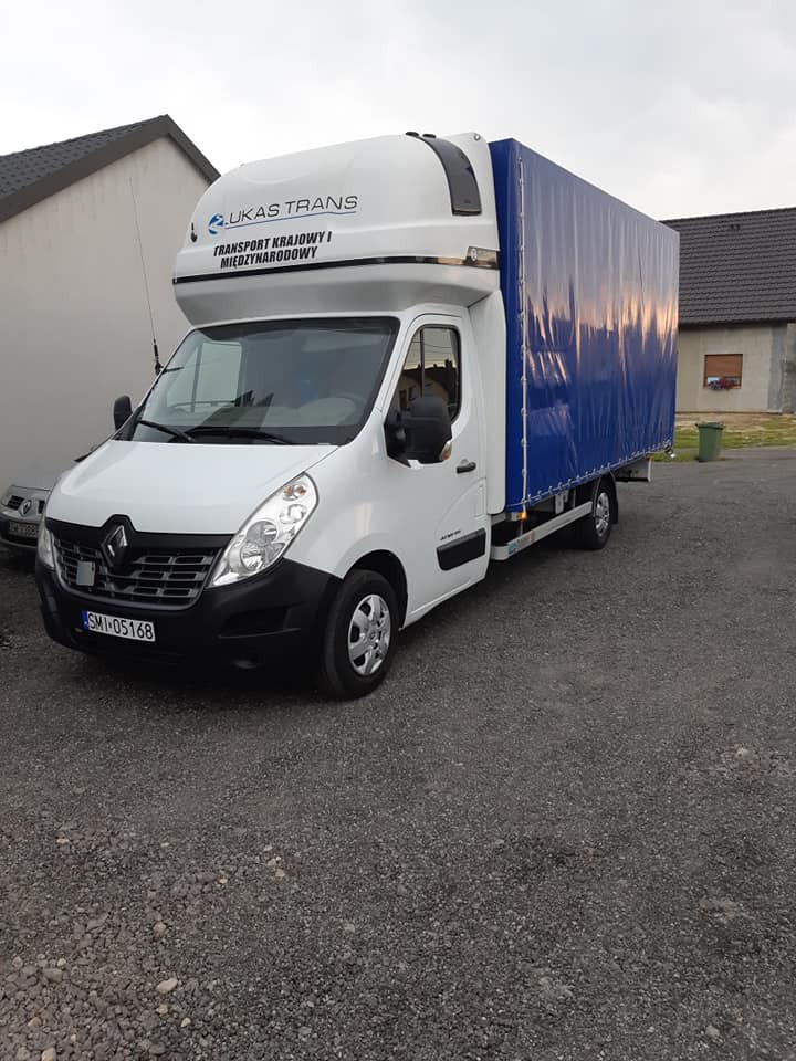 Biały bus Renault Master z plandeką w kolorze niebieskim, zaparkowany na żwirowym podłożu przed budynkami mieszkalnymi, z logo firmy 'Lukas Trans' na nadbudówce.