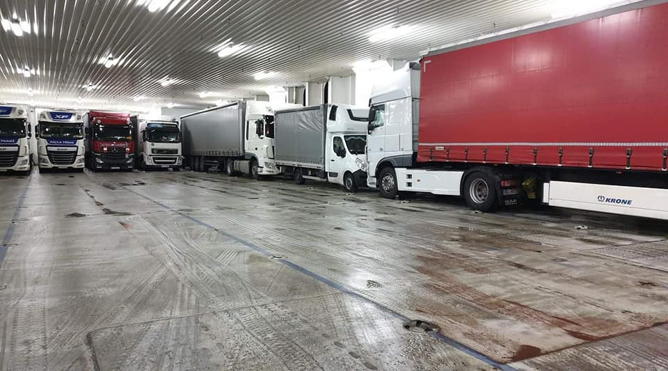 Ciężarówki różnych marek i rozmiarów ustawione w rzędzie wewnątrz promu lub magazynu, widoczne ślady opon na betonowej podłodze, oświetlenie sufitowe.