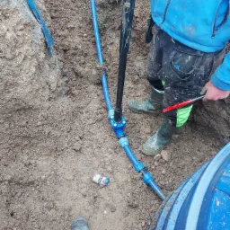 Montaż niebieskich rur w wykopie ziemnym, widoczne narzędzia i fragment zbiornika. Niedbale rzucona paczka papierosów na ziemi.