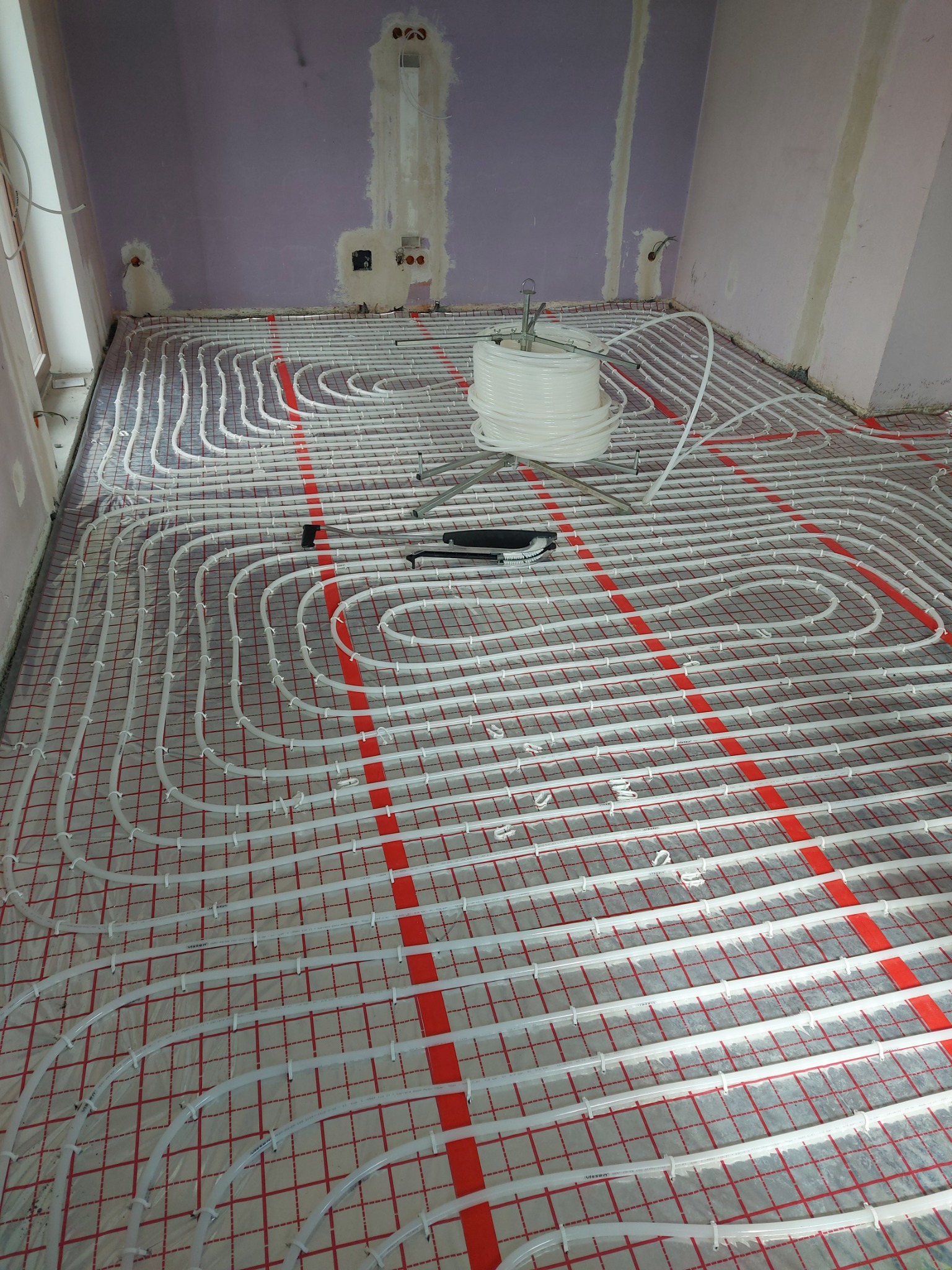 Białe rury ogrzewania podłogowego ułożone na czerwonej siatce montażowej w surowym pomieszczeniu z częściowo wykończonymi ścianami. Na podłodze stoi rolka z rurami i narzędzia.