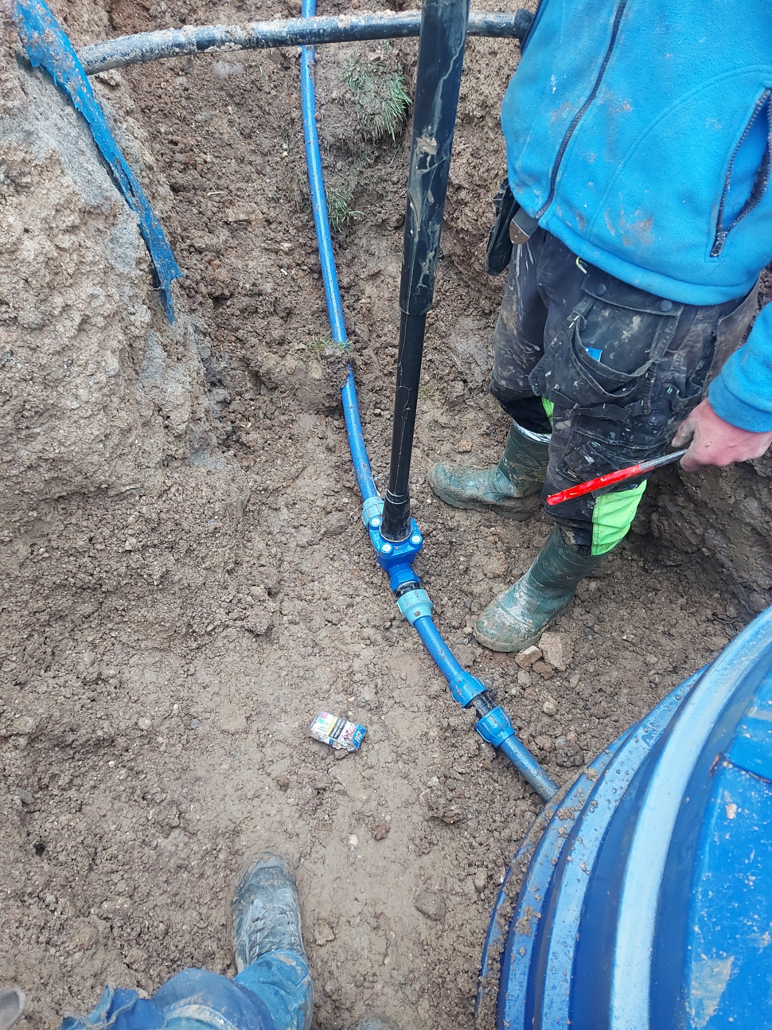 Montaż niebieskich rur w wykopie ziemnym, widoczne narzędzia i fragment zbiornika. Niedbale rzucona paczka papierosów na ziemi.