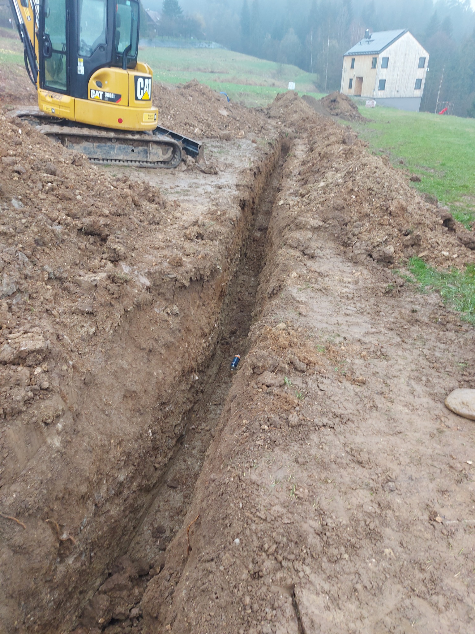 Żółta koparka CAT 305E2 na grząskim terenie obok wykopanego rowu, w tle budynek w stanie surowym zamkniętym i zalesione wzgórza we mgle.