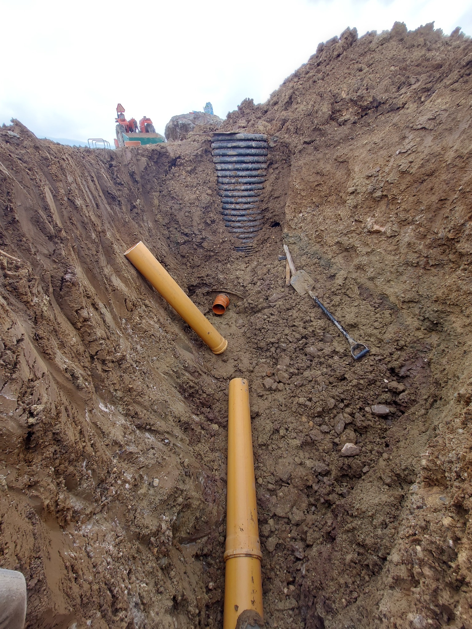 Głęboki wykop w ziemi z widocznymi rurami kanalizacyjnymi z tworzywa sztucznego, łopatą i sprzętem budowlanym w tle.