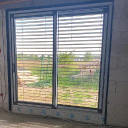 Zamknięte żaluzje fasadowe w oknie z czarną ramą, widok na pole i konstrukcję metalową na zewnątrz, surowy stan wykończenia wnętrza, widoczna folia ochronna z logo producenta na dolnej ramie okna.