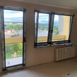 Montaż okien z widokiem na miasto, widoczne profile okienne zabezpieczone folią ochronną, grzejnik pod oknem, surowy stan wykończenia wnętrza.