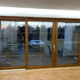 Przesuwne, trzyskrzydłowe drzwi balkonowe z brązową ramą i złotymi szprosami, widok na ogród o zmierzchu.