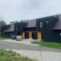 Nowoczesne segmenty bliźniacze z ciemną elewacją, brązowym dachem i drewnianymi bramami garażowymi, widok z zewnątrz na szarym brukowanym podjeździe, z zaparkowanym niebieskim samochodem i stertą...