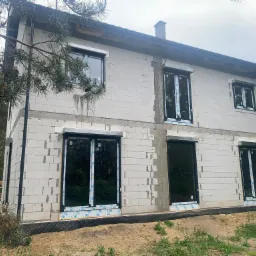 Surowy stan budynku w trakcie montażu stolarki okiennej i rolet zewnętrznych, widoczne bloczki betonowe, czarne ramy okienne i częściowo opuszczone rolety, zabezpieczenia folią na ramach, teren...