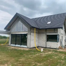 Surowy stan domu jednorodzinnego z zamontowanymi oknami z czarnymi ramami i dużym oknem trójkątnym na szczycie, widoczne żółte rury na zewnątrz, budynek w trakcie budowy, szara dachówka.