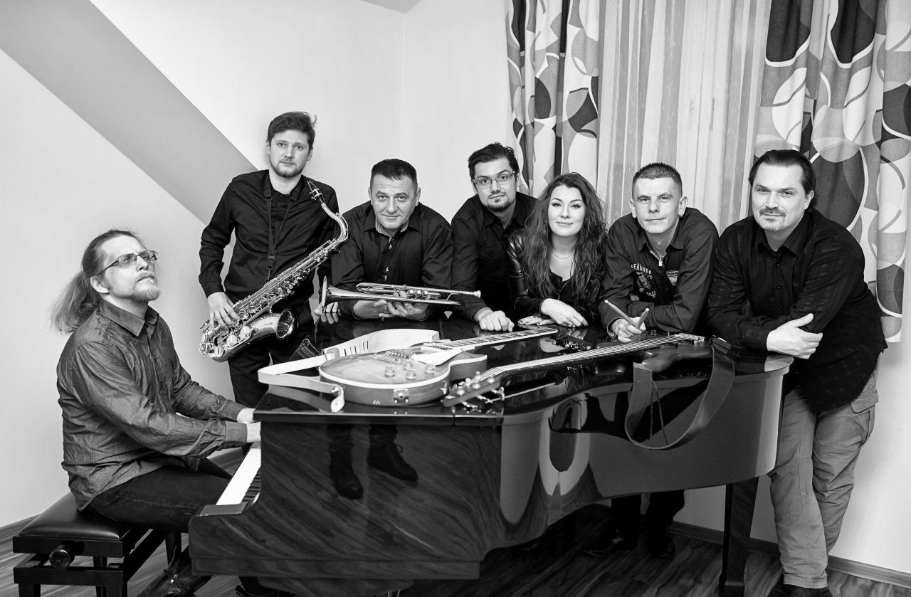 Czarno-białe zdjęcie siedmioosobowej grupy muzyków pozujących z instrumentami: saksofonem, trąbką, gitarami i fortepianem, na tle ściany i wzorzystej zasłony.