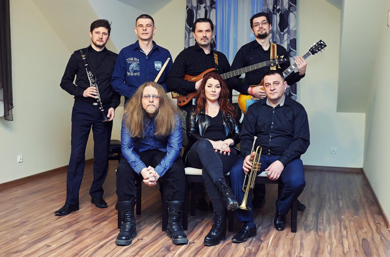 Siedmioosobowy zespół muzyczny w ciemnych strojach pozuje z instrumentami: klarnet, perkusja, gitara basowa, gitara elektryczna i trąbka.