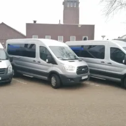 Trzy srebrne busy Ford Transit zaparkowane obok siebie na parkingu w pochmurny dzień, z budynkami w tle i widocznymi numerami rejestracyjnymi.