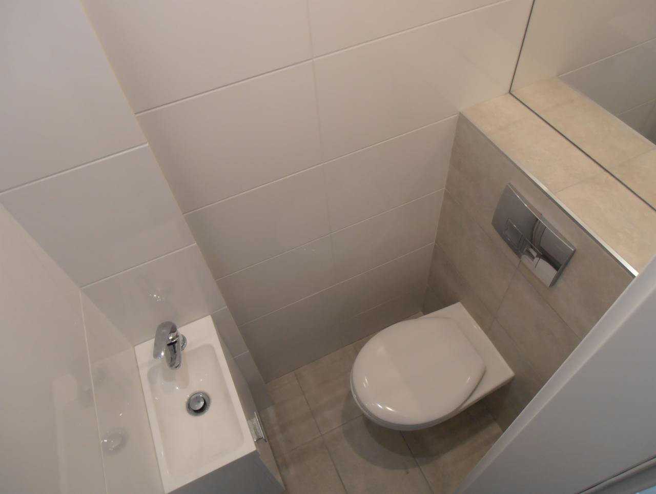 Nowoczesna, mała łazienka z wiszącą toaletą, umywalką narożną i dużym lustrem, wykończona białymi i beżowymi płytkami.