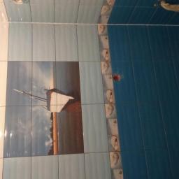 K-G Bud - Wykończona ściana łazienki z białymi, jasnoniebieskimi i ciemnoniebieskimi płytkami. Na jasnoniebieskich płytkach dekoracyjny motyw żaglówki na plaży.