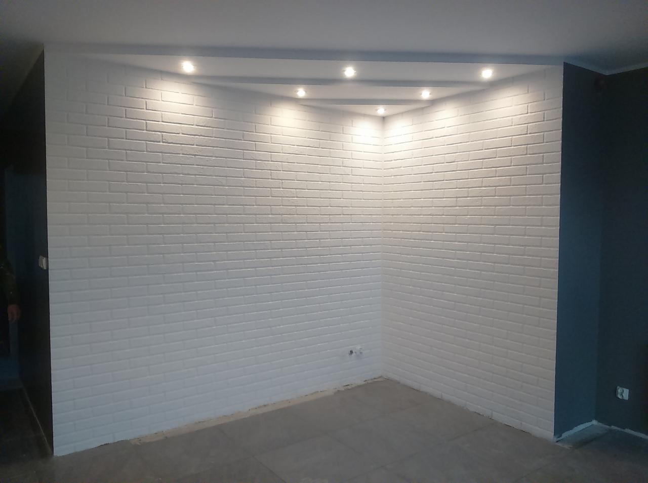 Wnętrze z białą ścianą wyłożoną cegiełkami, oświetlenie punktowe w suficie, szara podłoga, fragment ciemnej ściany w rogu.
