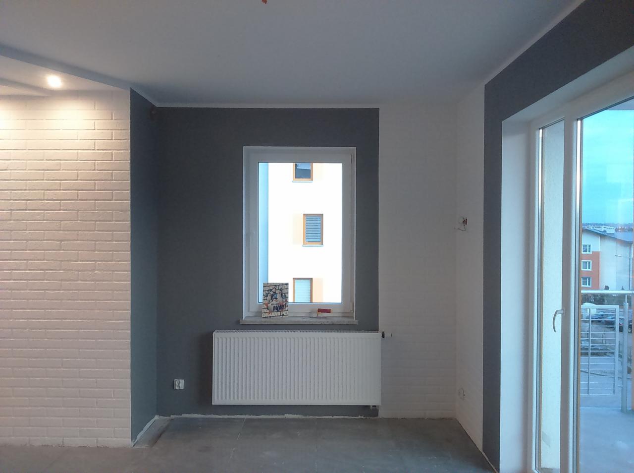 Wnętrze pokoju po malowaniu ścian na biało i szaro, z białą cegłą dekoracyjną na jednej ścianie, grzejnikiem pod oknem i widokiem na budynek zza okna.