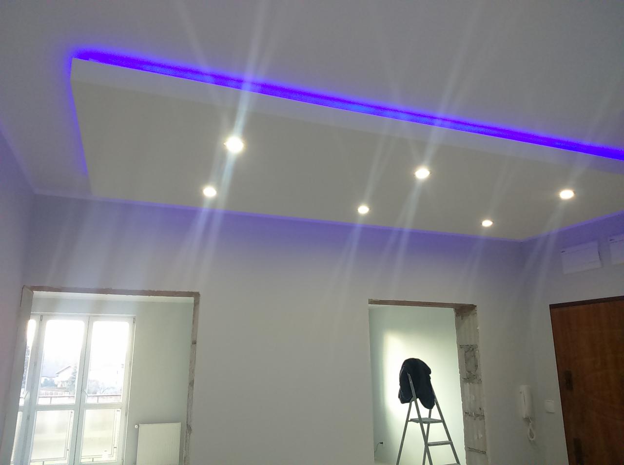 Nowoczesny sufit podwieszany z oświetleniem LED w kolorze fioletowym, widoczne otwory drzwiowe w stanie surowym oraz drabina z ubraniem, pokój w trakcie prac wykończeniowych.